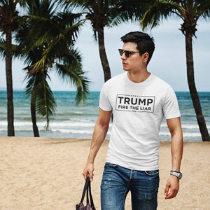 May include: A white t-shirt with black text that reads "TRUMP FIRE THE LIAR 2020". The person wearing the shirt is standing on a sandy beach with palm trees in the background.