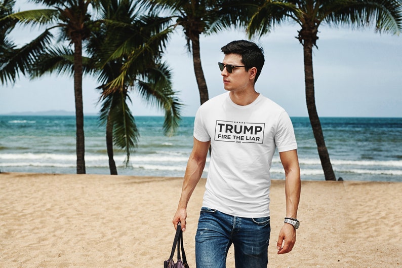 May include: A white t-shirt with black text that reads "TRUMP FIRE THE LIAR 2020". The person wearing the shirt is standing on a sandy beach with palm trees in the background.