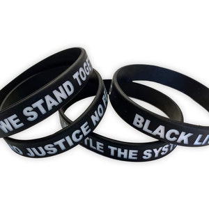 May include: Four black silicone wristbands with white lettering. The wristbands read "We Stand Together", "No Justice No Peace", "Black Lives Matter", and "Fight The System".
