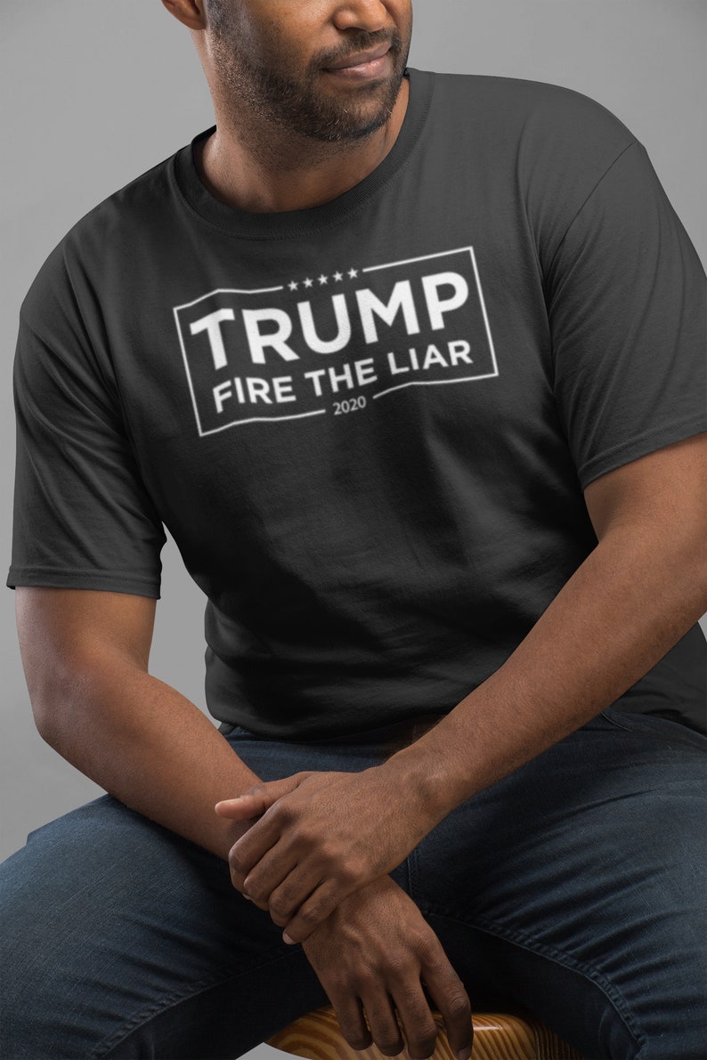 May include: A black t-shirt with a white rectangle that says "TRUMP FIRE THE LIAR - 2020" in all caps.