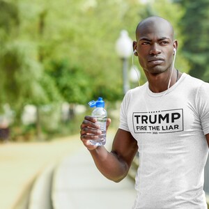 May include: A white t-shirt with a black rectangle on the front that says "TRUMP FIRE THE LIAR 2020". A person is wearing the shirt and holding a clear water bottle.