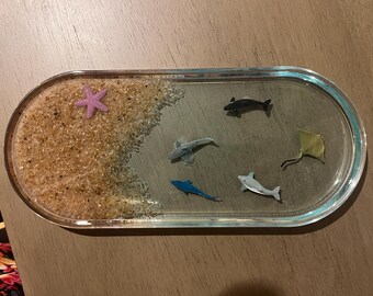 May include: An oval resin tray with a beach scene. The tray features a sandy area with a purple starfish and miniature sea creatures, including sharks, a stingray, and a dolphin. The tray has a clear, turquoise-colored border.