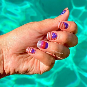 Peut inclure: Gros plan sur une main avec quatre ongles peints d'un vernis &agrave; ongles paillet&eacute; rouge, blanc et bleu avec un petit motif de drapeau am&eacute;ricain. La main repose sur une surface turquoise.