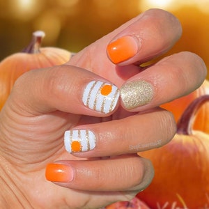 May include: A hand displaying a manicure with fall-themed nail art. The nails feature orange ombre, gold glitter, and white with gold striped designs, each with an orange dot. The background is a blurred image of pumpkins.