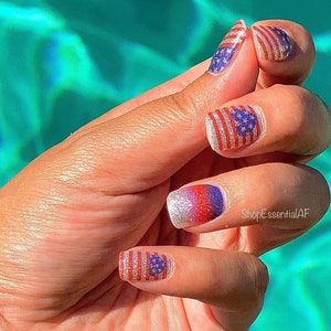 Peut inclure: Gros plan sur une main avec un nail art rouge, blanc et bleu repr&eacute;sentant des drapeaux am&eacute;ricains. Les ongles sont peints avec une base de paillettes argent&eacute;es et ont un effet ombr&eacute; rouge, blanc et bleu. Le texte "ShopEssentialAF" est visible sur la main.