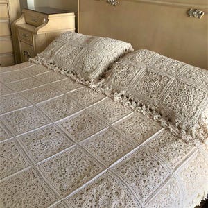 Könnte beinhalten: Eine cremefarbene, gehäkelte Tagesdecke und zwei passende Kissenbezüge. Die Tagesdecke besteht aus quadratischen Paneelen mit floralen und geometrischen Mustern. Die Kissenbezüge haben einen gewellten Rand. Das Bett befindet sich in einem Schlafzimmer mit cremefarbenem Kopfteil und Nachttisch.