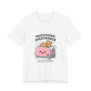 May include: White t-shirt featuring a cartoon toaster with two slices of bread. The toaster is pink with a smiling face and the text "PROFESSIONAL OVERTHINKER" and "LET'S GET THIS BREAD (AND THEN WORRY ABOUT IT)".