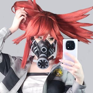 May include: A person wearing a black and gray gas mask, red wig, and a gray jacket with black stripes. The person is holding a white smartphone. The jacket has a yellow and black emblem.