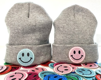May include: Two gray knit beanies with a smiley face patch. The patches are light blue and pink. A collection of glittery smiley face patches in various colors are scattered below the beanies.