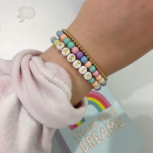 Peut inclure: Un bracelet à perles multicolores avec le nom "NASIK" écrit en lettres dorées. Le bracelet est porté au poignet d'une personne. Le bracelet est posé sur un livre avec le texte "CHASE YOUR DREAMS" imprimé dessus.