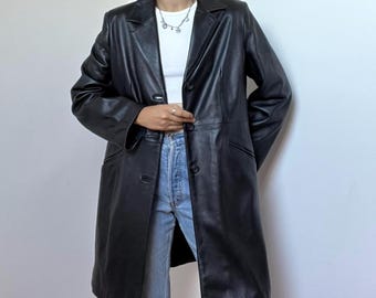 May include: A black leather coat featuring a notched lapel, long sleeves, and a button-front closure. The coat is worn over a white top and light blue denim jeans. The design includes two side pockets and a structured fit that reaches mid-thigh.