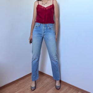 May include: A deep red satin camisole featuring a delicate lace trim along the neckline and thin spaghetti straps, paired with light-wash blue denim jeans that have a classic straight-leg cut and a standard button-fly closure.