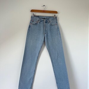 May include: Light blue denim jeans hanging on a wooden hanger against a white wall.