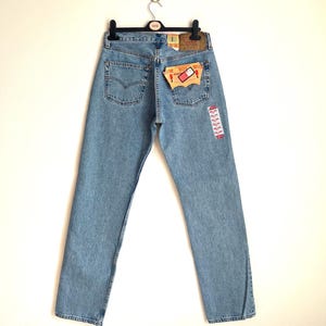 May include: Light blue denim jeans hanging on a black hanger. The jeans have a classic five-pocket design, a leather patch, and a red and white tag. The jeans are a light wash and appear to be new.