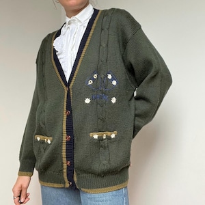 May include: Dark green cable-knit cardigan with a V-neck and button closure. The sweater features navy blue and gold trim, along with embroidered floral details. The cardigan has two front pockets and a relaxed fit.