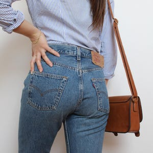 May include: A light blue and white striped long-sleeve button-down shirt paired with medium-wash blue denim jeans featuring a tan leather patch on the back waistband and a small red tab on the back pocket, accompanied by a brown leather crossbody bag.