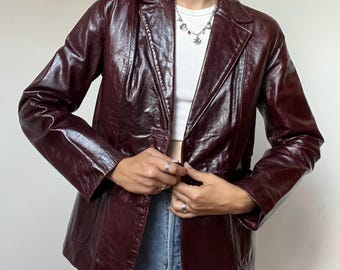 Può includere: Un blazer bordeaux scuro con una texture lucida e stropicciata, caratterizzato da revers classici, maniche lunghe e chiusura con bottoni. Il blazer è indossato sopra una canotta bianca a coste e jeans blu, abbinato a una collana con ciondoli in argento.