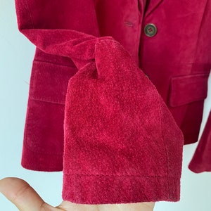 K&ouml;nnte beinhalten: Eine Nahaufnahme eines burgunderfarbenen Wildleder-Blazers mit einem einzigen Knopfverschluss. Der Blazer hat einen klassischen Stil und ist aus einem weichen, luxuri&ouml;sen Stoff gefertigt.