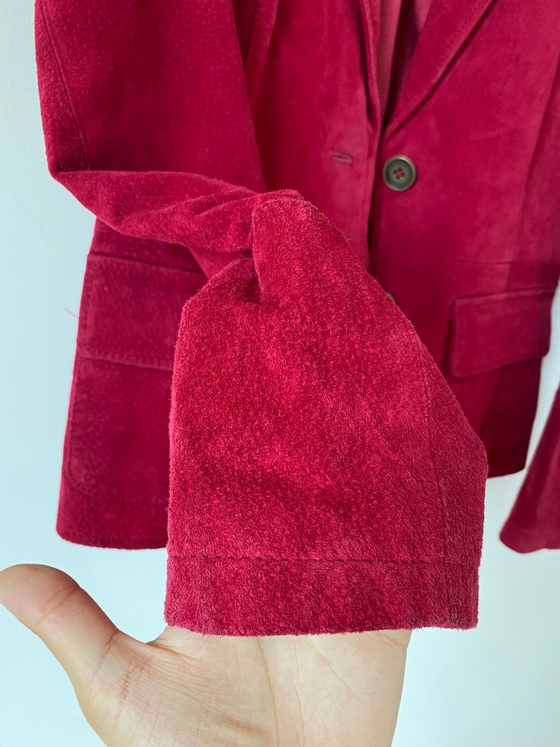 K&ouml;nnte beinhalten: Eine Nahaufnahme eines burgunderfarbenen Wildleder-Blazers mit einem einzigen Knopfverschluss. Der Blazer hat einen klassischen Stil und ist aus einem weichen, luxuri&ouml;sen Stoff gefertigt.
