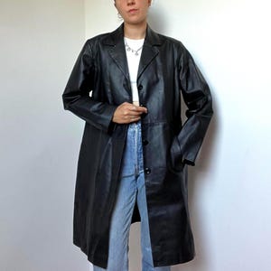 May include: A black long-line coat featuring a notched lapel collar, long sleeves, and a front button closure. The coat is worn over a white cropped top and light blue denim jeans. The garment has a smooth finish and reaches mid-thigh length.