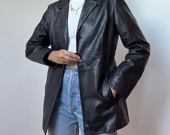 Könnte beinhalten: Ein schwarzer Lederblazer mit steigendem Revers, langen Ärmeln und einer Knopfleiste auf der Vorderseite. Die Jacke wird über einem weißen T-Shirt mit Rundhalsausschnitt und einer hellblauen Jeans getragen. Eine silberfarbene Kette ist sichtbar.