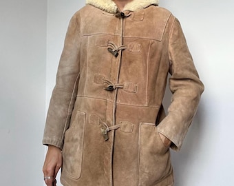 May include: A tan suede duffle coat with a hood and toggle closures. The coat has a light-colored faux fur lining around the hood and two front pockets. The coat is a medium length.