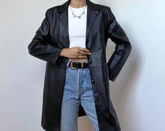 May include: A black leather coat featuring a notched collar, long sleeves, and a button-front closure. The coat is worn over a white crew-neck top and light blue denim jeans with a black belt featuring a gold-toned buckle.