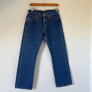 May include: A pair of medium-wash blue denim jeans featuring a classic five-pocket design, a button-fly closure, and copper-toned rivets. The jeans have a straight-leg cut and are displayed hanging from a wooden hanger against a plain wall.
