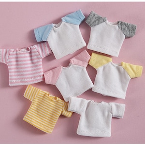 Op de afbeelding: Een set van tien miniatuur poppenhemden in verschillende kleuren en patronen, waaronder roze en wit gestreept, geel en wit gestreept, wit met blauwe mouwen, wit met grijze mouwen en wit met gele mouwen.