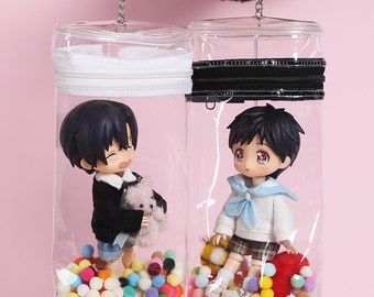 May include: Two clear plastic zippered pouches with colorful pom poms inside. Each pouch contains a small doll with a different outfit and a plush toy. The pouches have a silver chain attached to the top.