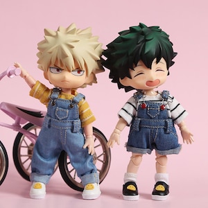 May include: Two small dolls wearing denim overalls. One doll has blonde hair and is wearing a yellow striped shirt. The other doll has green hair and is wearing a white and black striped shirt. Both dolls are wearing white socks and black shoes with yellow smiley faces.