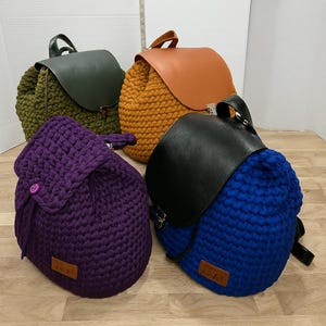 May include: Four handmade crochet backpacks in various colors: purple, green, orange, and blue. Each backpack features a flap closure and a leather top. The backpacks are displayed on a light-colored wooden surface.