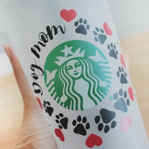 May include: White reusable cup with a green Starbucks logo and the text "dog mom" in black. The cup is decorated with black paw prints and red hearts.