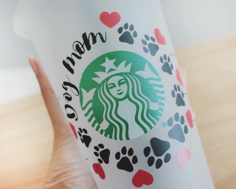 May include: White reusable cup with a green Starbucks logo and the text "dog mom" in black. The cup is decorated with black paw prints and red hearts.