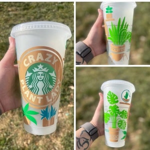 May include: A white reusable cup with a gold and green design. The cup has the text "Crazy Plant Lady" on it. The design includes a green plant with a brown pot and a green leaf.