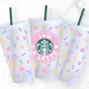 K&ouml;nnte beinhalten: Drei durchsichtige Plastikbecher mit Deckeln und Strohhalmen. Die Becher haben ein rosa-wei&szlig;es Design mit Herzen, Sternen und Streuseln. Der mittlere Becher hat einen rosa Kreis mit dem Text "I love a latte" und ein gr&uuml;nes Starbucks-Logo.