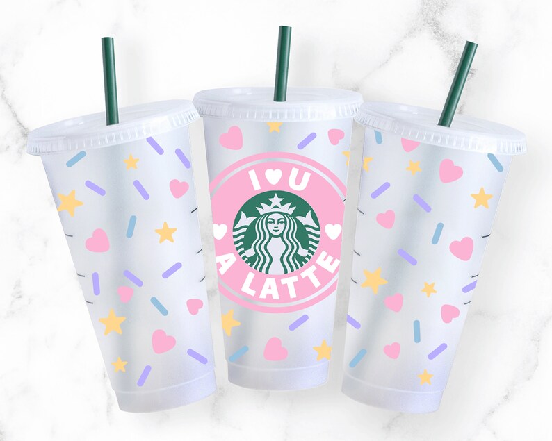 K&ouml;nnte beinhalten: Drei durchsichtige Plastikbecher mit Deckeln und Strohhalmen. Die Becher haben ein rosa-wei&szlig;es Design mit Herzen, Sternen und Streuseln. Der mittlere Becher hat einen rosa Kreis mit dem Text "I love a latte" und ein gr&uuml;nes Starbucks-Logo.