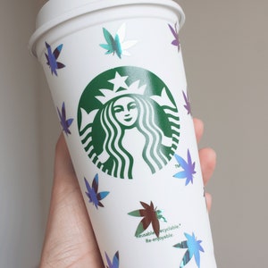 Peut inclure: Gobelet à café blanc réutilisable avec un logo vert et des décalcomanies de feuilles de marijuana iridescentes. Le gobelet a un couvercle blanc et le texte "Reusable recyclable Re-enjoyable" imprimé sur le côté.