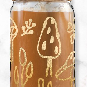 May include: A glass jar with a wooden lid and a metal straw. The jar is filled with a brown liquid and has a gold design of mushrooms and leaves.