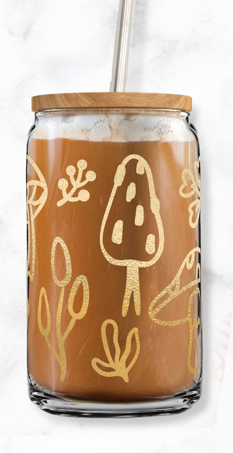 May include: A glass jar with a wooden lid and a metal straw. The jar is filled with a brown liquid and has a gold design of mushrooms and leaves.