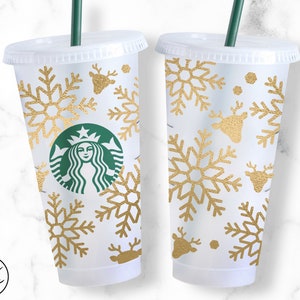 Puede incluir: Dos vasos de plástico transparentes con diseños de copos de nieve y renos dorados. Los vasos tienen tapas y pajitas. El logotipo de Starbucks está en uno de los vasos.