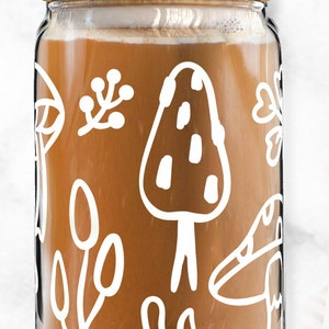 May include: A glass jar with a wooden lid and a clear glass straw. The jar is filled with a brown liquid and has a white design of mushrooms, leaves, and other nature-inspired elements.