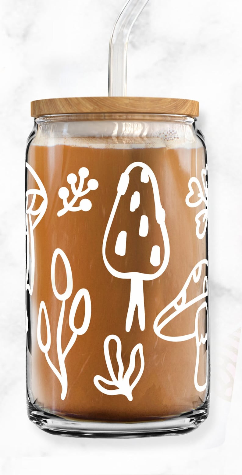 May include: A glass jar with a wooden lid and a clear glass straw. The jar is filled with a brown liquid and has a white design of mushrooms, leaves, and other nature-inspired elements.
