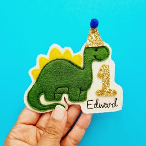 May include: A green felt dinosaur with a yellow spine and a gold glitter party hat. The number "1" and the name "Edward" are also in gold glitter. The dinosaur is on a white background.