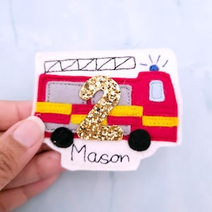 May include: A red, yellow, grey and black felt fire engine applique with a gold glitter number 2. The name "Mason" is written in black script below the truck. The applique is on a white background.