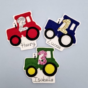 May include: Three felt tractor-shaped birthday decorations. One is red with a silver glitter number 2 and the name "Henry". Another is blue with a gold glitter number 1 and the name "Lucas". The third is green with a pink glitter number 3 and the name "Isabella".