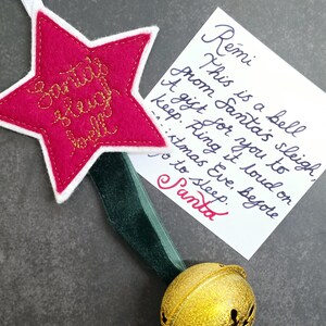 May include: A gold jingle bell attached to a dark green velvet ribbon and a pink felt star ornament with white trim. The star is embroidered with the text Santa's sleigh bell. A white card features handwritten text addressed to Remi from Santa.
