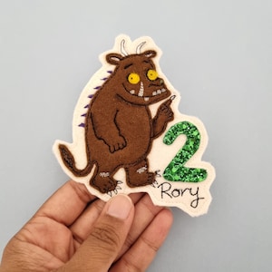 May include: A brown felt applique featuring a cartoon monster with yellow eyes and a green glitter number 2. The name "Rory" is written below the number. The applique is on a cream-colored background.