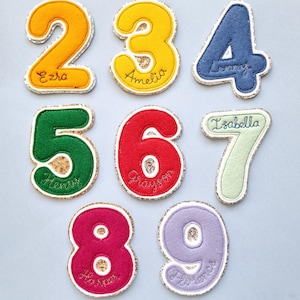 May include: A selection of colourful felt number patches with a glittery gold border. Numbers 2 to 9 are shown in various colours, including yellow, blue, green, red, pink, and purple. Each number has a name embroidered on it.