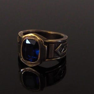 May include: A gold-toned ring with a large, square-cut, deep blue gemstone. The ring has a thick band with geometric designs. The gemstone is set in a bezel setting, and the ring is displayed on a reflective black surface.
